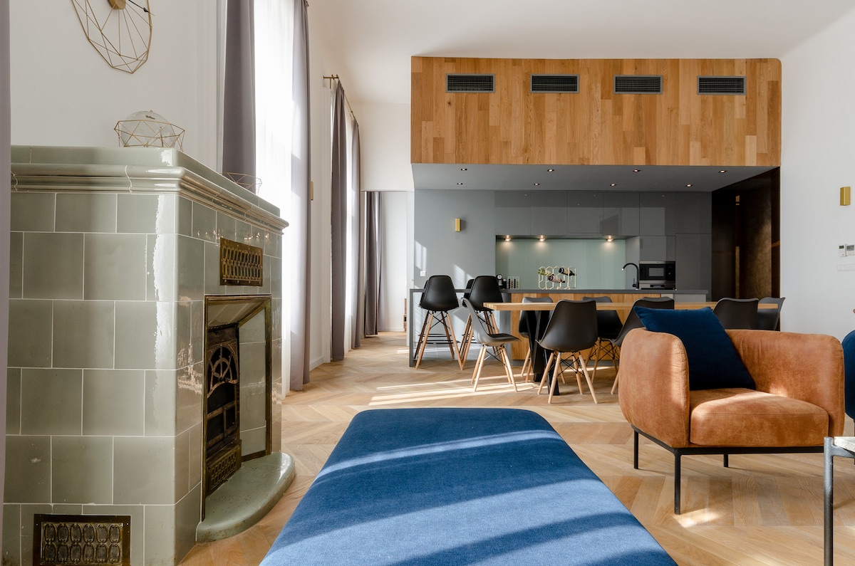 A spacious living area showcases a custom-designed couch and a large-screen TV, complemented by a vintage fireplace. The open-plan kitchen is equipped with modern appliances. Light floods through the tall windows, enhancing the warmth of the wooden accents and stylish furnishings.