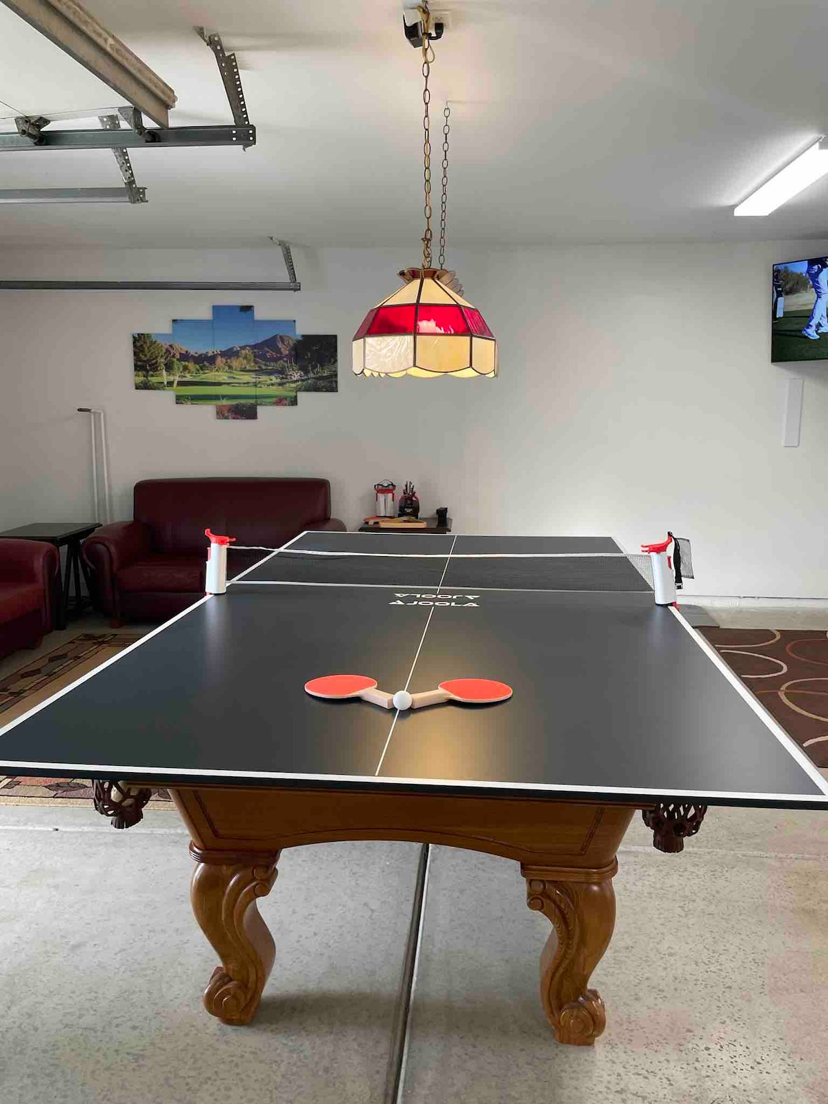 A multi-functional game table is featured, set up for ping pong, with two paddles and a ball positioned on the surface. The space is illuminated by a vintage-style pendant light, and a seating area with two maroon sofas is visible in the background.