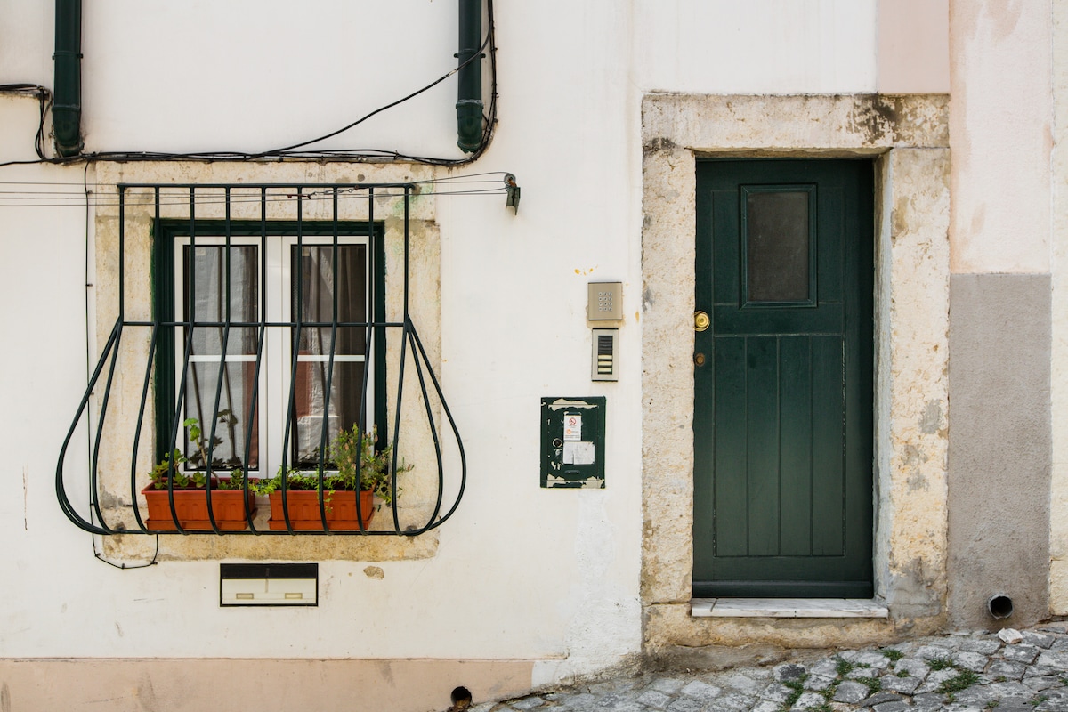 A traditional façade is highlighted by a green door and a window adorned with wrought iron bars. The window box features potted plants, adding a touch of greenery to the crisp white exterior. The stone wall exhibits wear, contributing to the building's character.