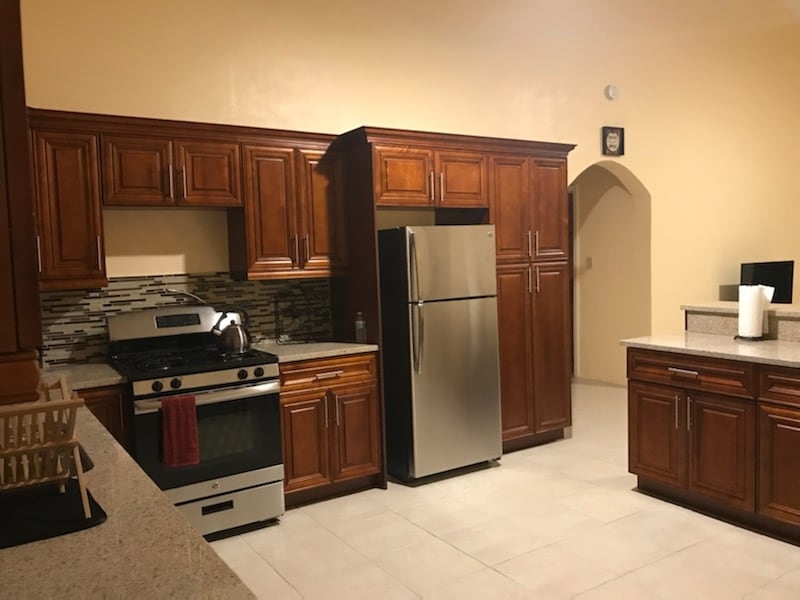 A warm-toned kitchen features ample wooden cabinetry and stainless steel appliances, including a refrigerator and a gas stove. Countertops are made of light-colored stone, and a backsplash of varied tiles adds subtle texture. Natural light floods in, highlighting the spacious layout.