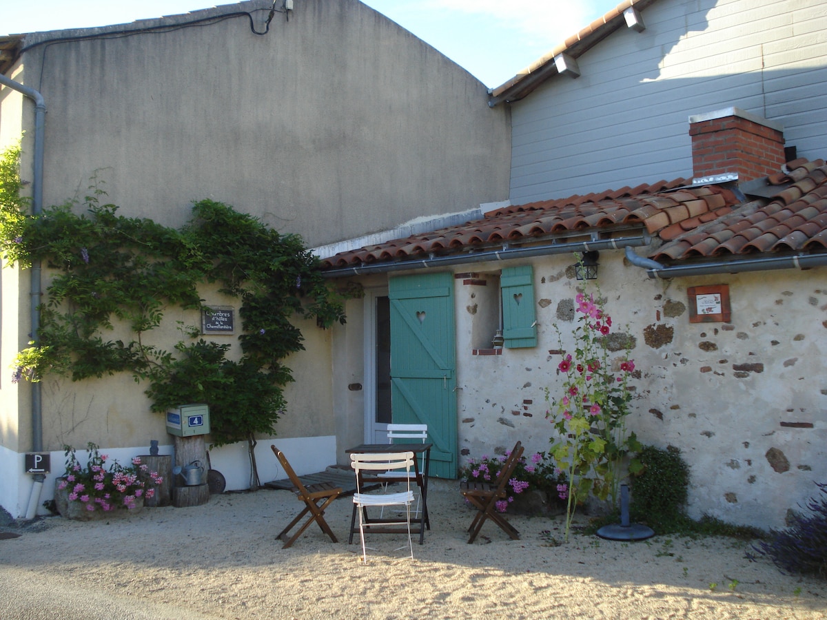 An outdoor area is present, featuring a sandy surface with a few scattered stones. There are small, wooden tables and chairs positioned thoughtfully amongst flowering plants. A rustic building, characterized by stone walls and a tiled roof, stands in the background, complemented by a green door.