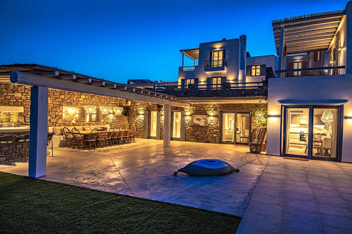 An outdoor area is illuminated by soft lighting in the evening, highlighting a spacious terrace with a dining table and chairs. A lounge area is visible, alongside a small pool. The structure incorporates stone walls and modern architecture, blending into the serene nighttime setting.