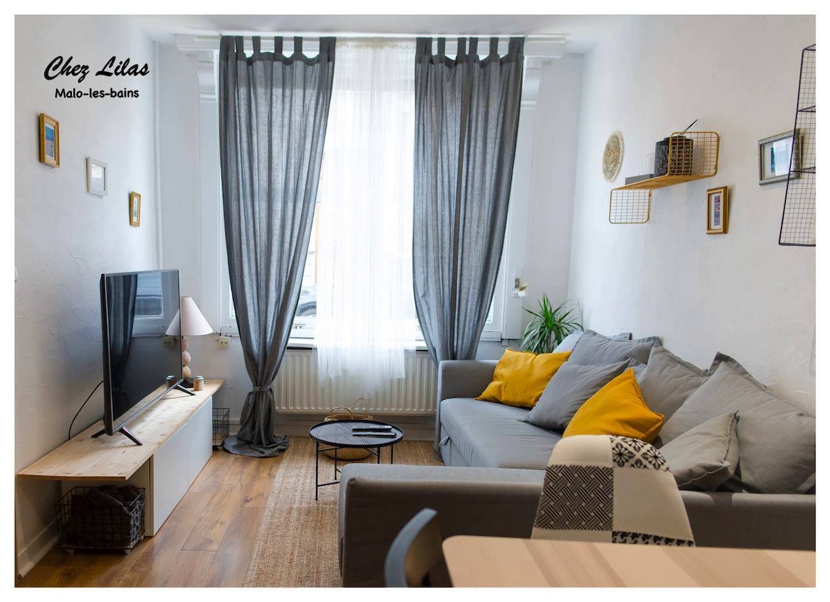 A bright living area is displayed, featuring a comfortable gray sofa adorned with yellow and patterned cushions. Large sheer curtains frame a window that allows natural light to fill the room. A television is mounted on a light wood media console beside a circular coffee table.