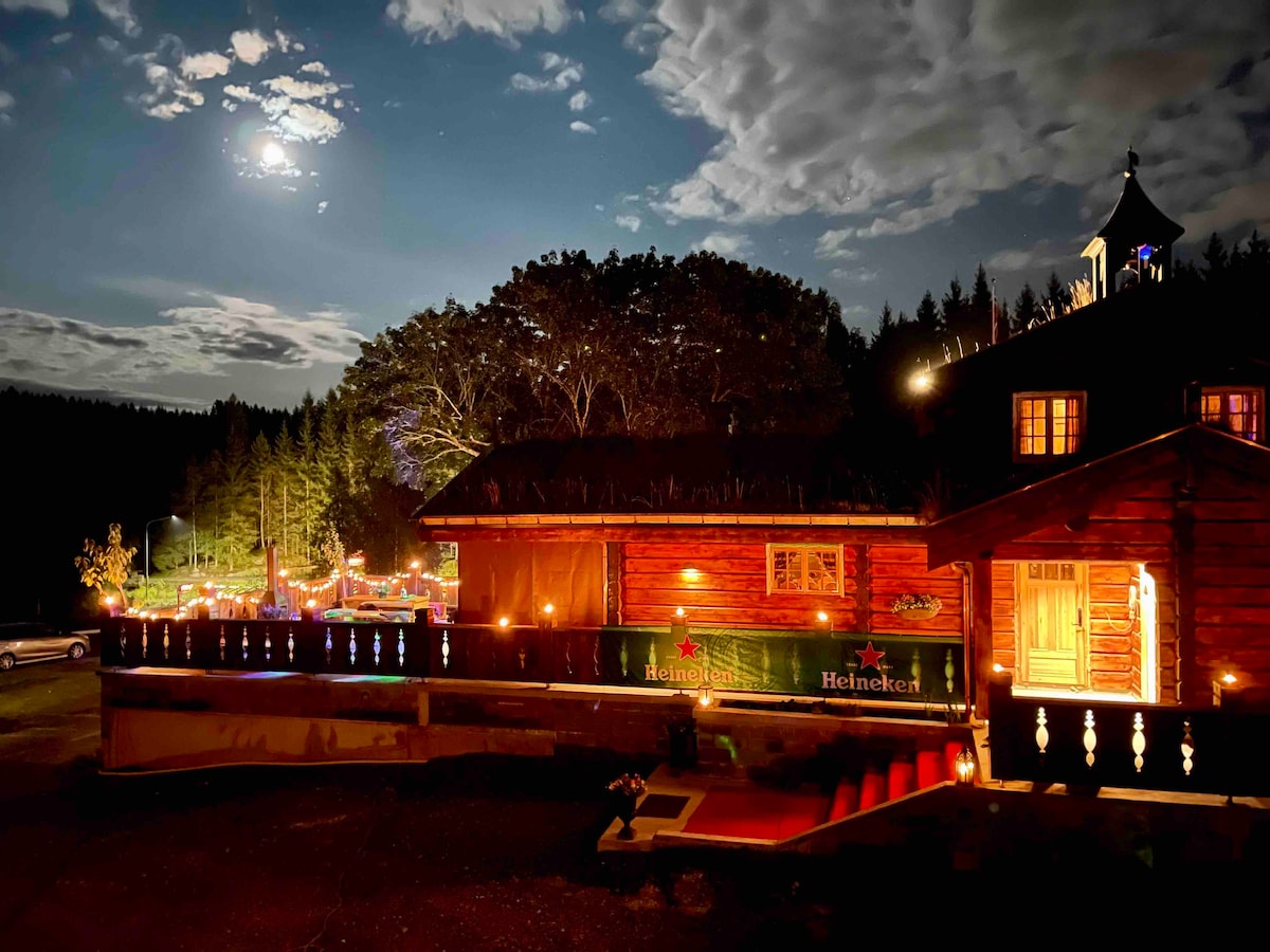 A newly built wooden cabin is illuminated under a moonlit sky. Soft lighting enhances the structure, with a forest backdrop creating a serene atmosphere. The entrance is marked by welcoming open doors, inviting guests to explore the cozy interior.