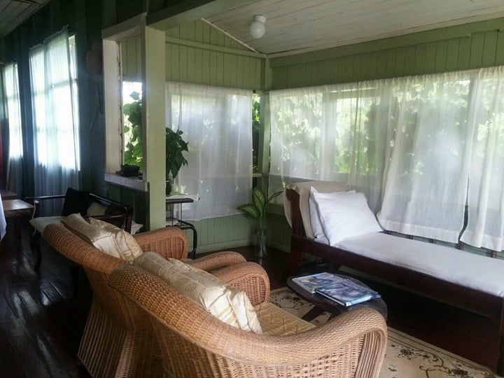 An airy lounge area features two wicker chairs and a daybed, highlighted by sheer white curtains allowing soft natural light. A small table holds magazines, while potted plants add a touch of greenery to the welcoming setting.