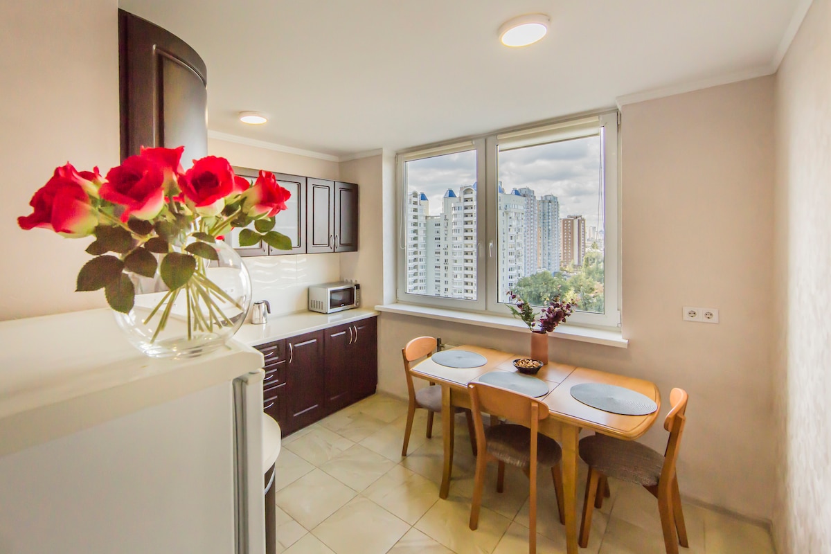 A well-appointed kitchen is visible, featuring modern cabinetry and a dining area with a round table and four chairs. A vase with red roses adds a touch of color. Large windows provide a view of the city skyline, allowing natural light to brighten the space.