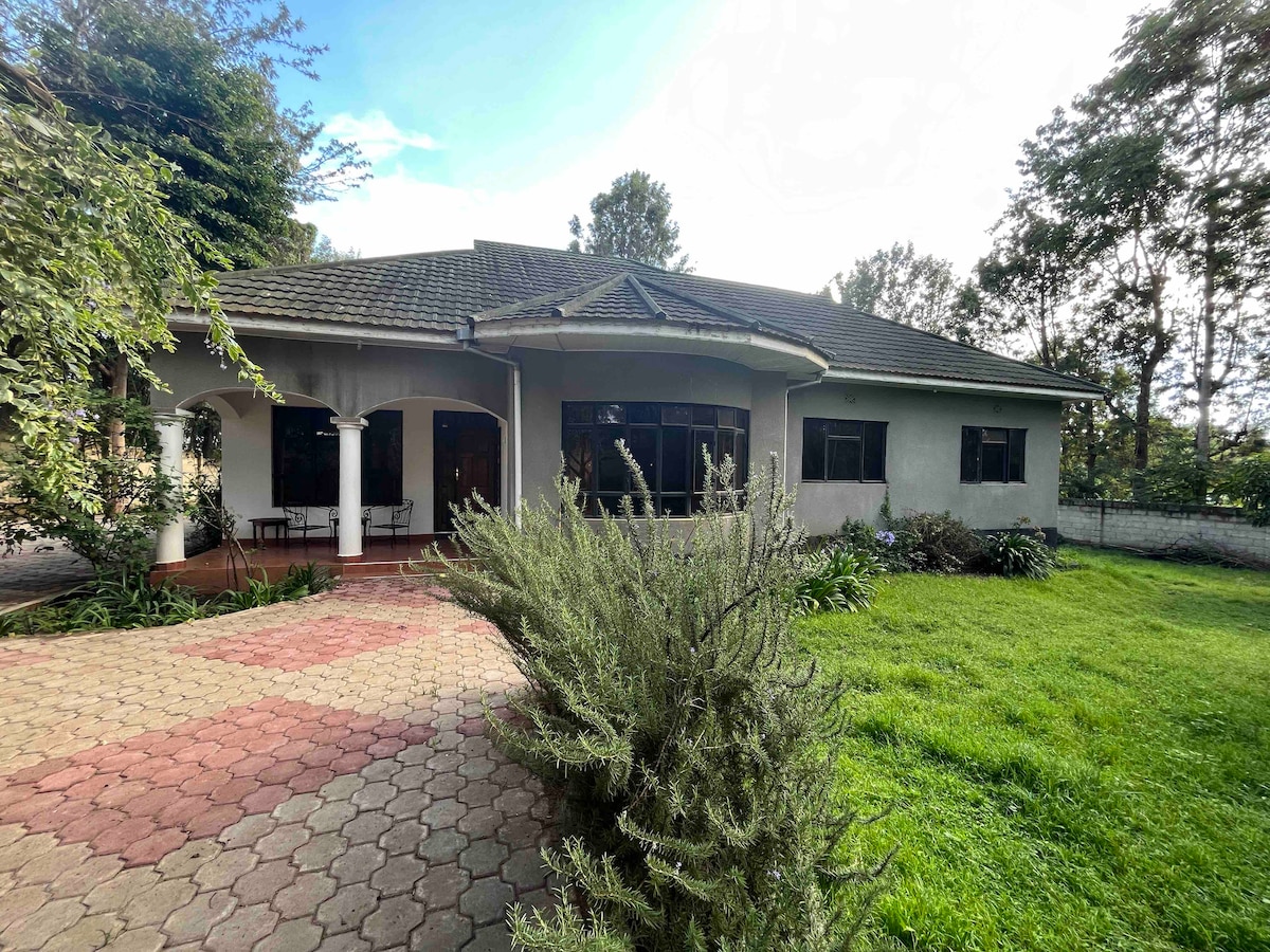 A spacious single-story lodge is surrounded by well-maintained gardens, featuring a large front porch supported by columns. The lawn is adorned with greenery, and a patterned stone pathway leads to the entrance. Large windows allow natural light to illuminate the interior.