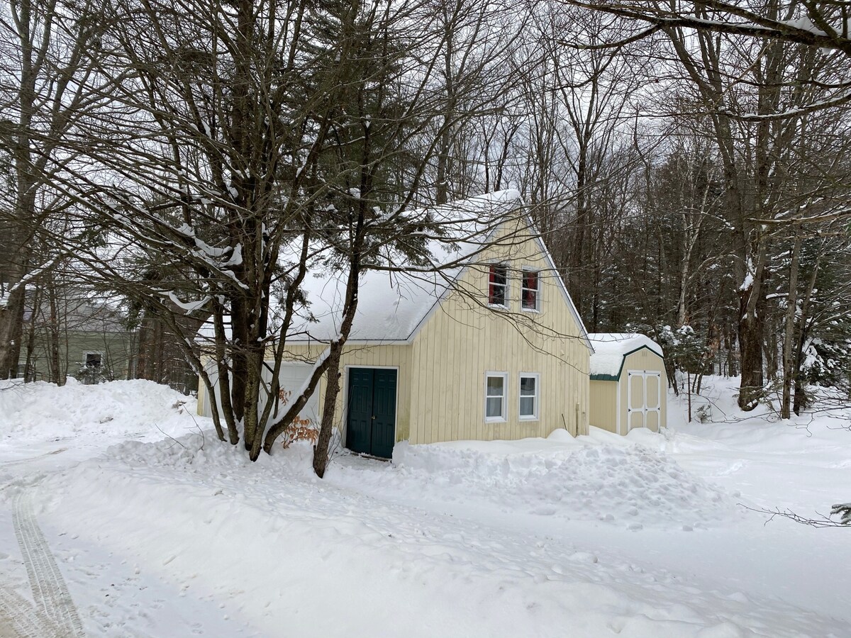 A charming yellow house is set among snow-covered trees, showcasing a steep roof and multiple windows. A small shed is visible beside the house, adding to the serene winter landscape. Snow blankets the ground, creating a peaceful atmosphere.