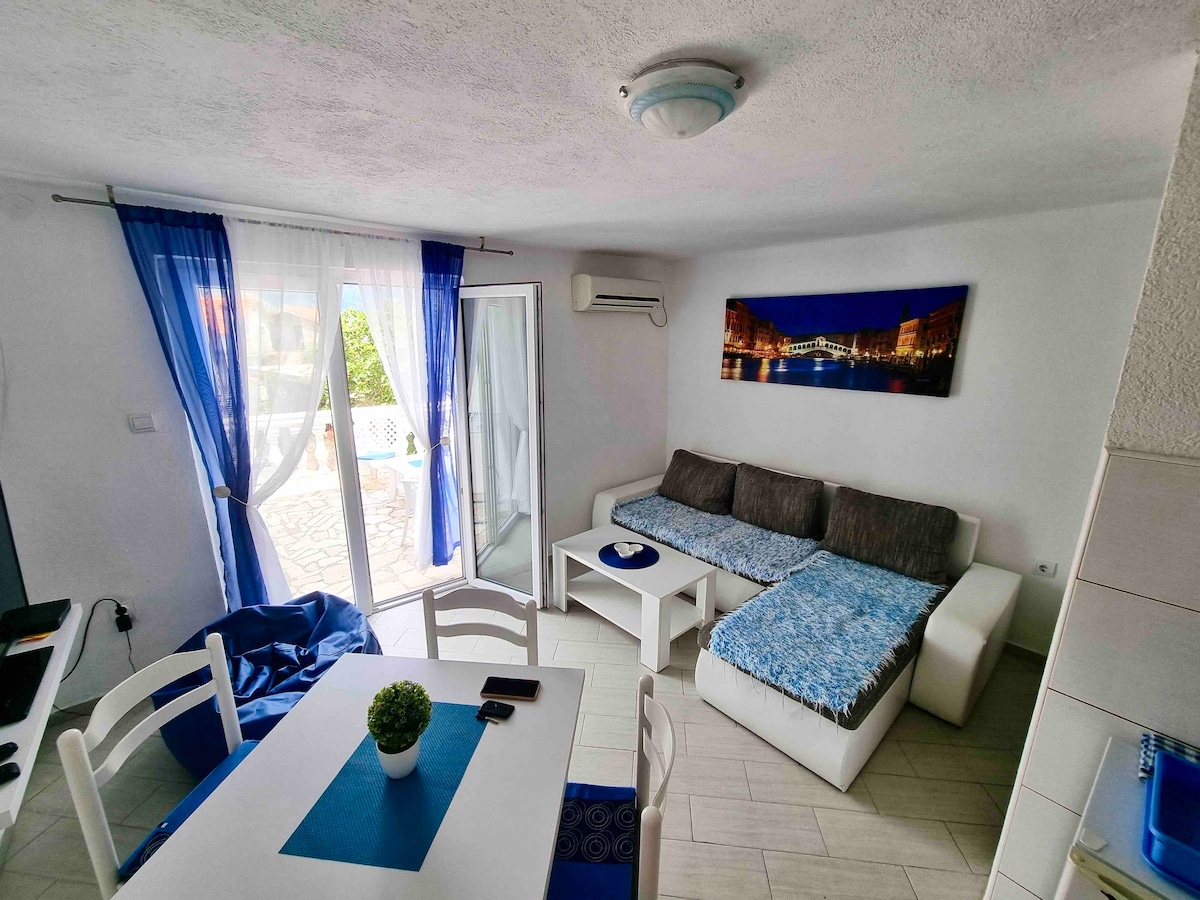 A bright living area is illustrated with a combination of a white sectional sofa and a coffee table. Sheer curtains frame a glass door that leads to the balcony. A small dining table with white chairs is set alongside a decorative plant, enhancing the room's inviting atmosphere.