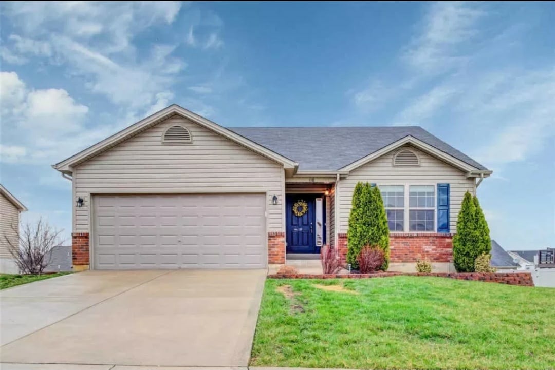 A single-story house features a light-colored exterior with a brick accent. The front porch is framed by two evergreen shrubs, and a well-maintained lawn leads up to the concrete driveway. A welcoming door is adorned with a decorative wreath.