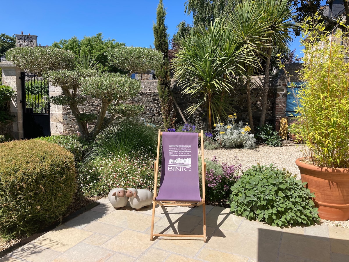 A serene garden area is presented, featuring a comfortable lounge chair and vibrant greenery. Various plants add texture, while decorative stones provide a natural pathway. A glimpse of blue can be seen in the background, enhancing the tranquil outdoor space.