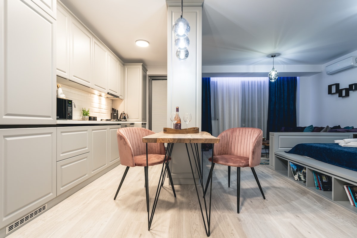 A modern dining area is featured, with a wooden table flanked by two pink chairs. The kitchen cabinetry provides a clean, cohesive look, while light fixtures offer gentle illumination. A cozy resting area is visible in the background, complemented by soft lighting and draped curtains.
