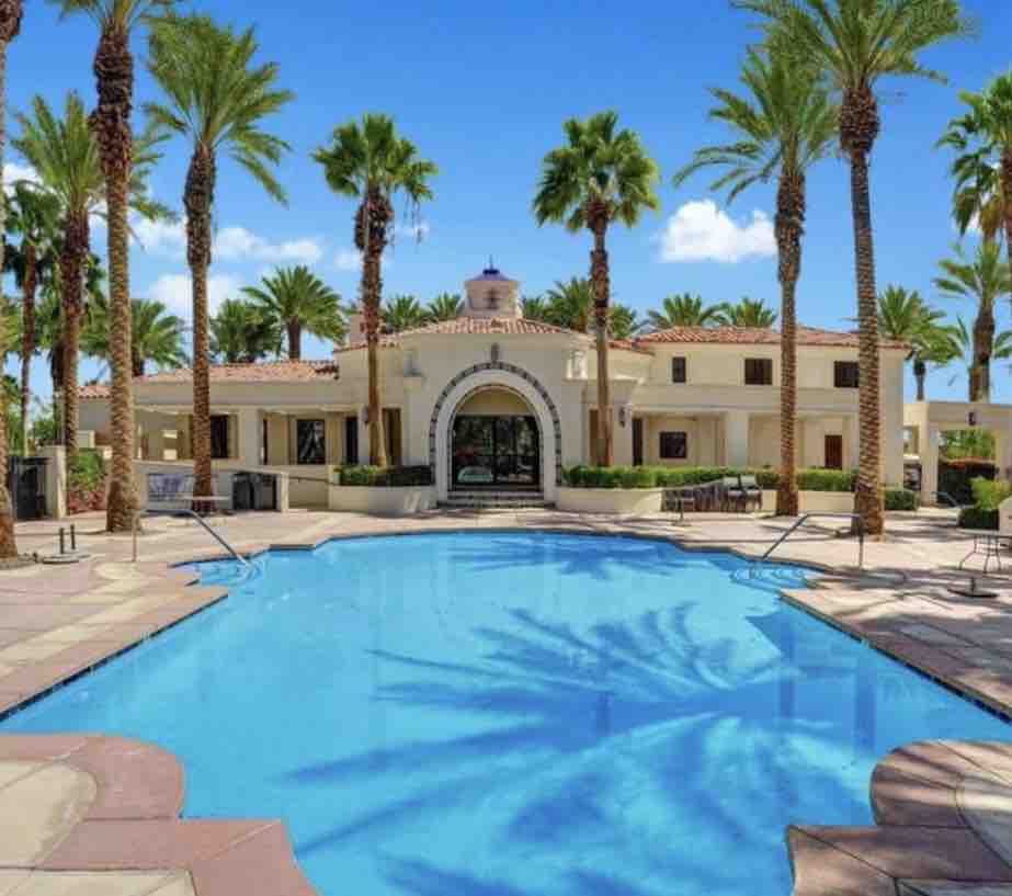 A resort-style clubhouse is set beside a large, inviting pool, framed by swaying palm trees. The building features a distinctive architectural arch, providing a welcoming entrance with vibrant blue skies overhead, reflecting a serene atmosphere for relaxation and leisure.