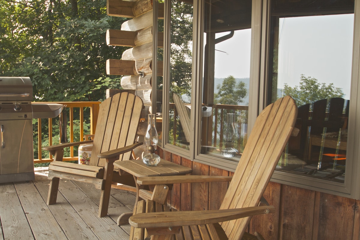 A wooden deck features two Adirondack chairs positioned beside a small table, creating a comfortable seating area. A gas grill is situated nearby, and large windows offer a view of the surrounding trees and the outdoor landscape.