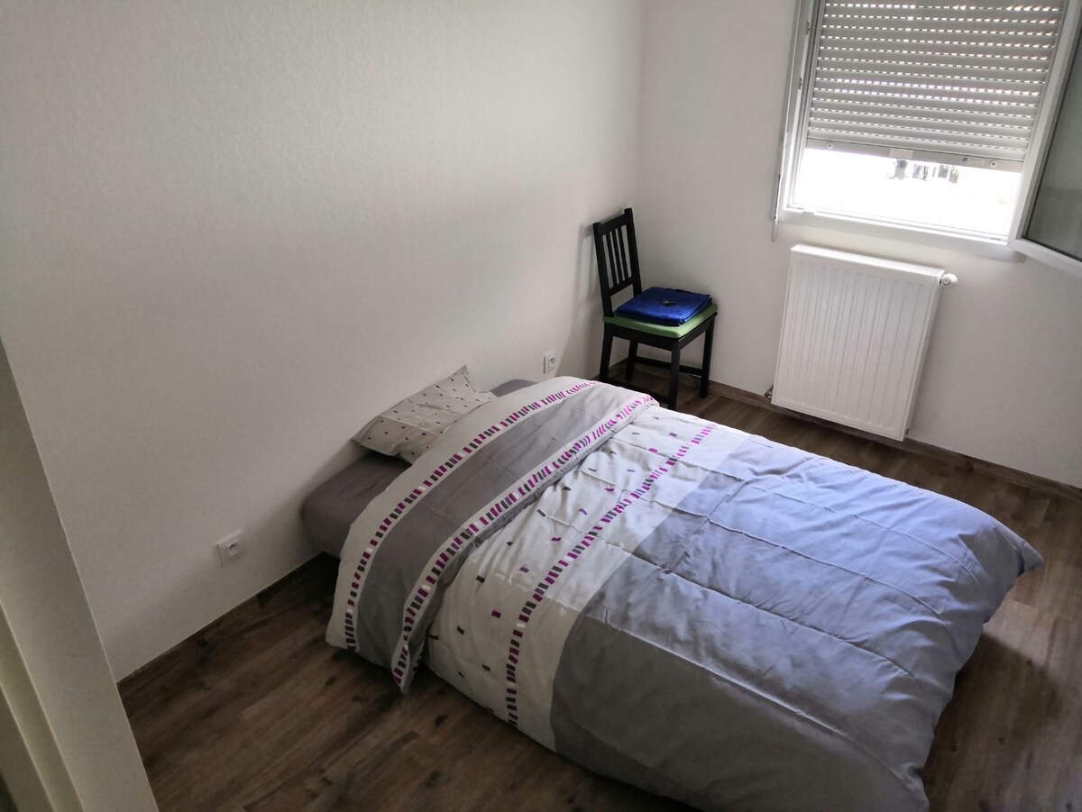 A simple sleeping area features a low bed with a light grey comforter and decorative bedding. A chair sits against the wall beside a window, which is covered by blinds, allowing for controlled natural light. A radiator is located near the window.
