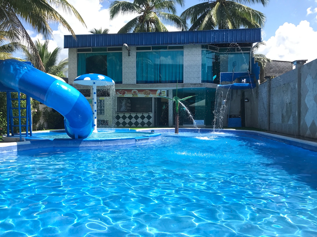A vibrant swimming pool area features a large blue water slide alongside a children's play area. Water cascades down from the structure, creating a refreshing ambiance. Lush palm trees provide shade, and the building's glass facade reflects the clear sky, enhancing the lively atmosphere.