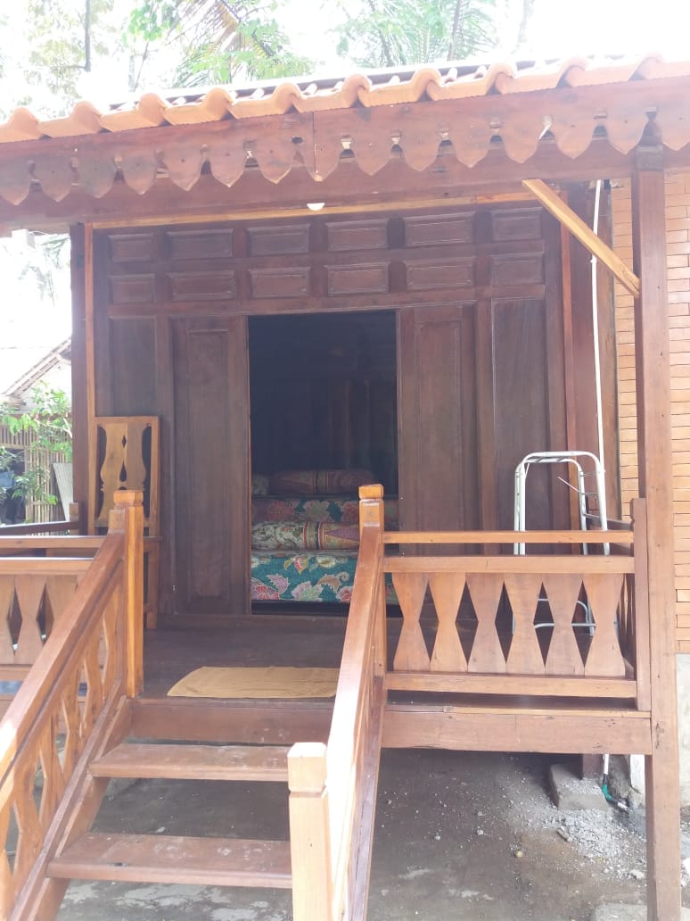 The exterior of a wooden structure is visible, showcasing intricately carved details. Steps lead up to a covered porch, where vibrant patterned bedding can be seen through an open doorway, inviting guests into the cozy interior.