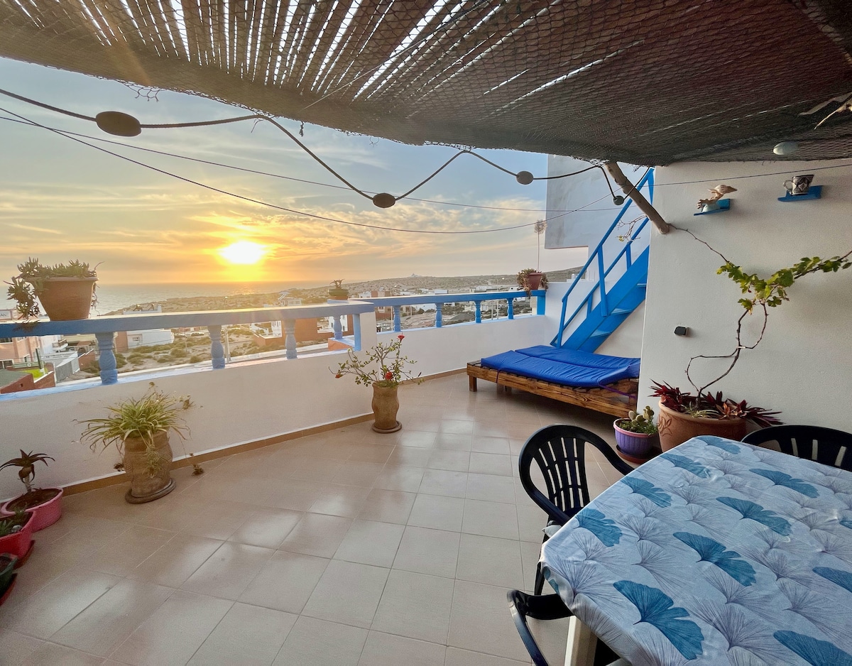 A spacious roof-top terrace is visible, featuring seating arrangements and potted plants. A large blue mat lies on the floor. The scene includes a sunset view over the horizon, with warm colors illuminating the sky, complemented by decorative elements along the walls.