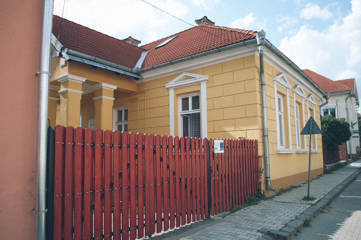 The exterior of the guesthouse is highlighted by yellow walls and a red wooden fence. The building features multiple windows with decorative white trim, and a tiled roof. A sidewalk runs alongside the house, offering a glimpse of the surrounding neighborhood.
