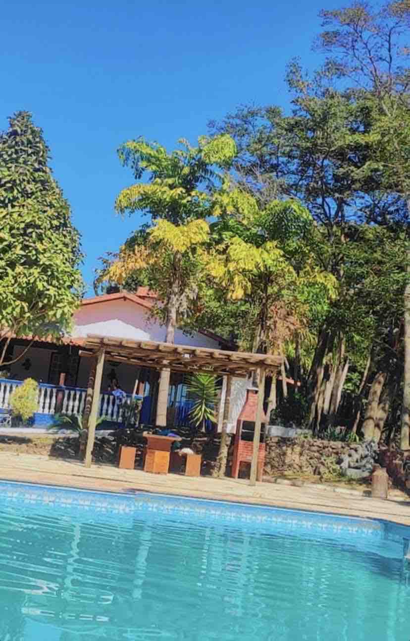 A serene view of a swimming pool with clear blue water reflects the bright sky. Lush greenery surrounds the area, complemented by a rustic wooden pergola in the foreground. The house in the background is partially obscured by trees, creating a tranquil atmosphere.