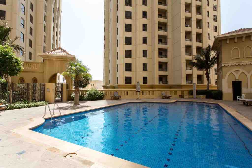 A swimming pool is centrally located within a landscaped area, bordered by tall buildings. Palm trees are positioned around the pool, which features clear blue waters and is equipped with a set of steps for easy access.