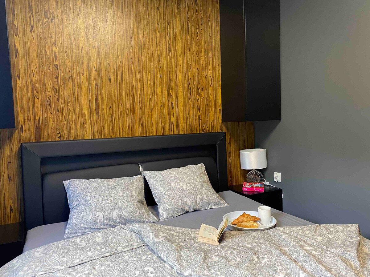 A well-appointed bedroom features a dark wooden accent wall alongside a sleek black headboard. Soft bedding is complemented by a stylish bedside table with a lamp. A book and a cup of coffee with a croissant are arranged invitingly on the bed.