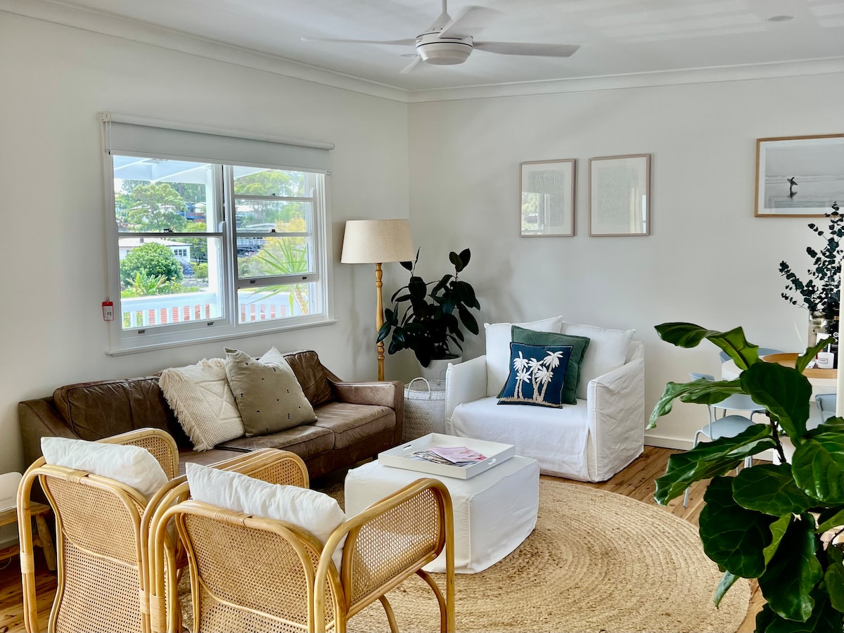 A welcoming living area features a mix of comfortable seating, including a leather sofa and white armchairs. A round jute rug anchors the space, while large windows allow natural light to fill the room. Potted plants and framed art add a touch of warmth.