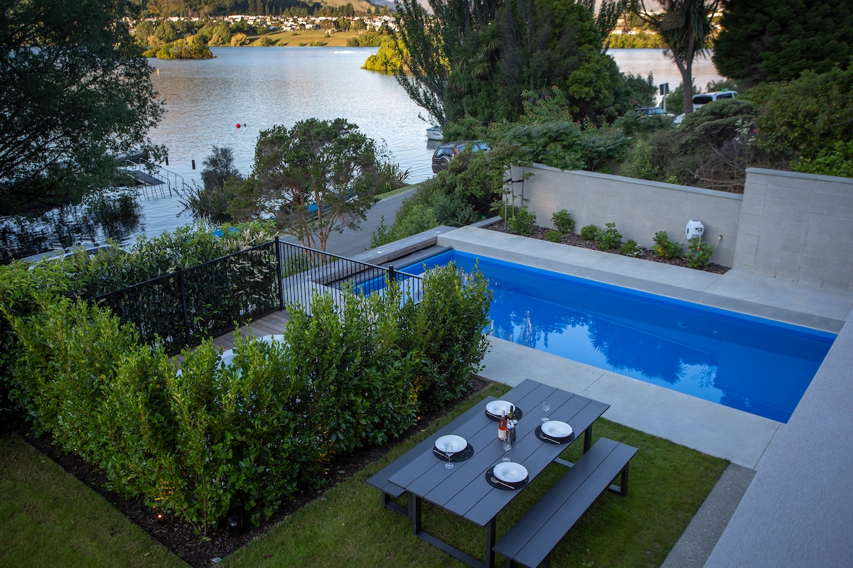 An outdoor space features a heated pool bordered by neatly trimmed shrubs and grass. A dining table with four place settings is positioned nearby, providing a view of the lake and surrounding trees. The tranquil setting offers a peaceful atmosphere.