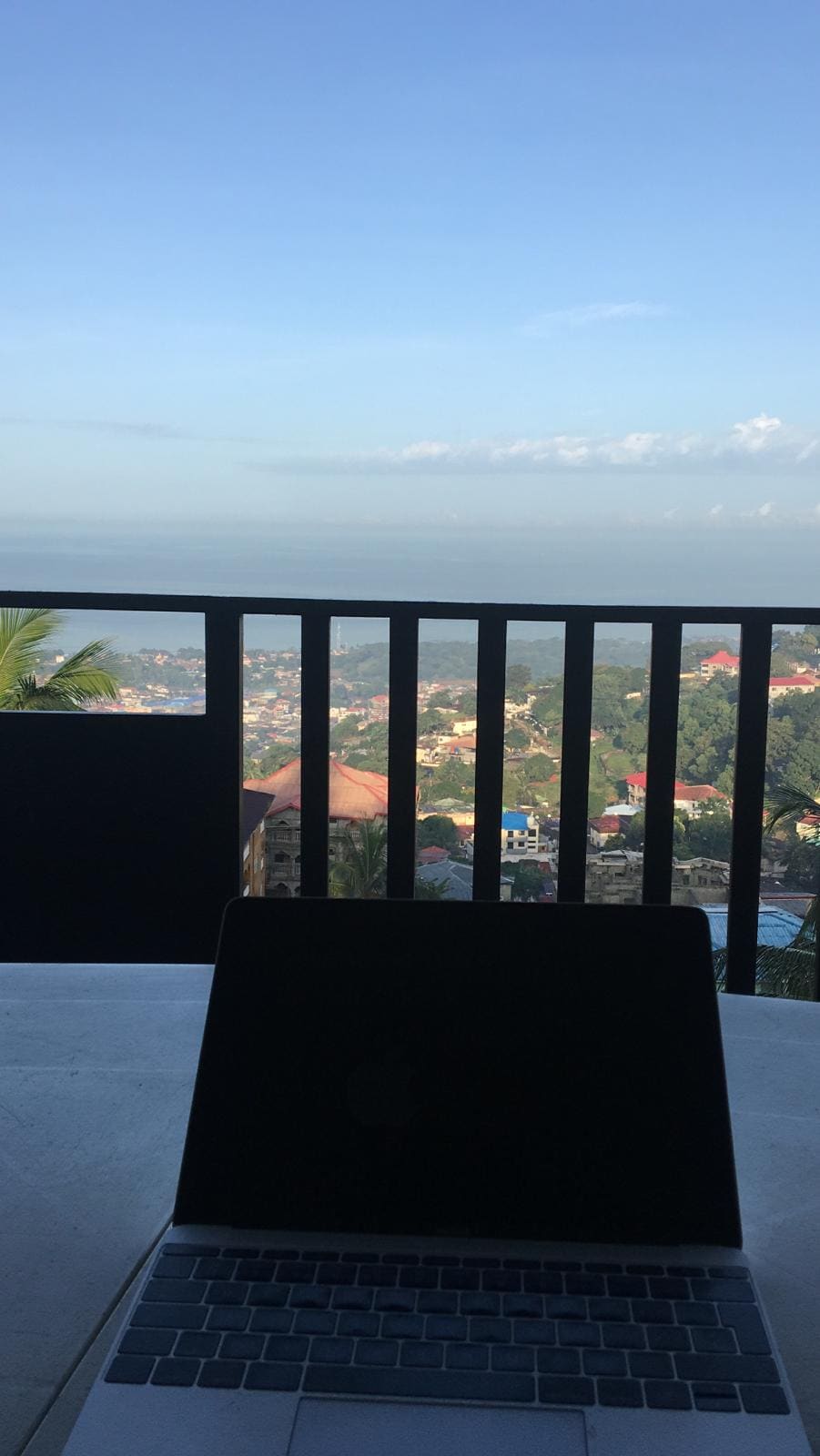 A view of the Atlantic Ocean is presented from a balcony. The railing's dark design frames the scene, while a table is visible in the foreground, holding a laptop. The distant landscape shows a mix of greenery and buildings under a clear blue sky.
