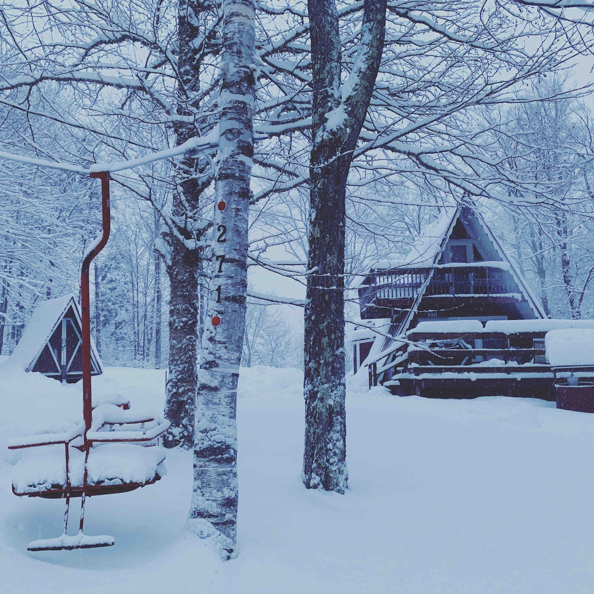 A snow-covered landscape features an A-Frame cabin surrounded by tall trees. A rustic ski lift chair rests beside a birch tree, both blanketed in snow. The tranquil scene conveys a sense of winter solitude, with soft snowflakes gently covering the ground.