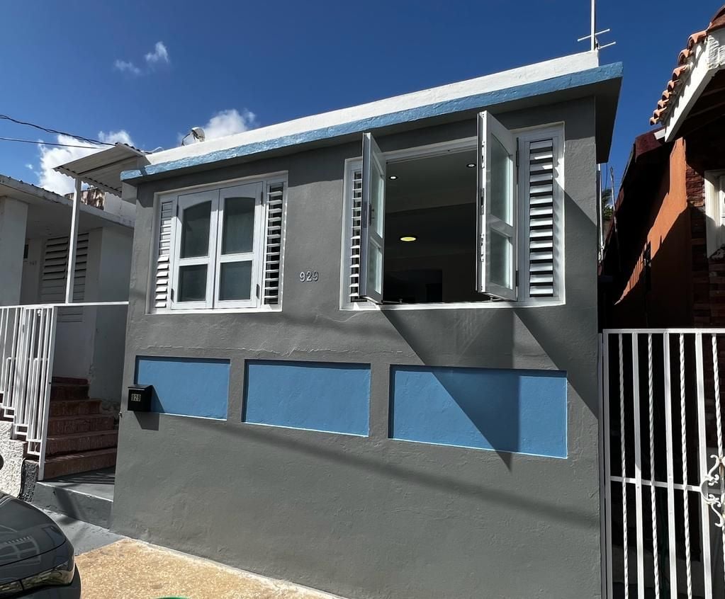 A small beach house is displayed with multiple windows opened, providing a breezy atmosphere. The exterior features a combination of gray and blue paint with white trim. Lush greenery is visible nearby, enhancing the inviting entrance to the home.