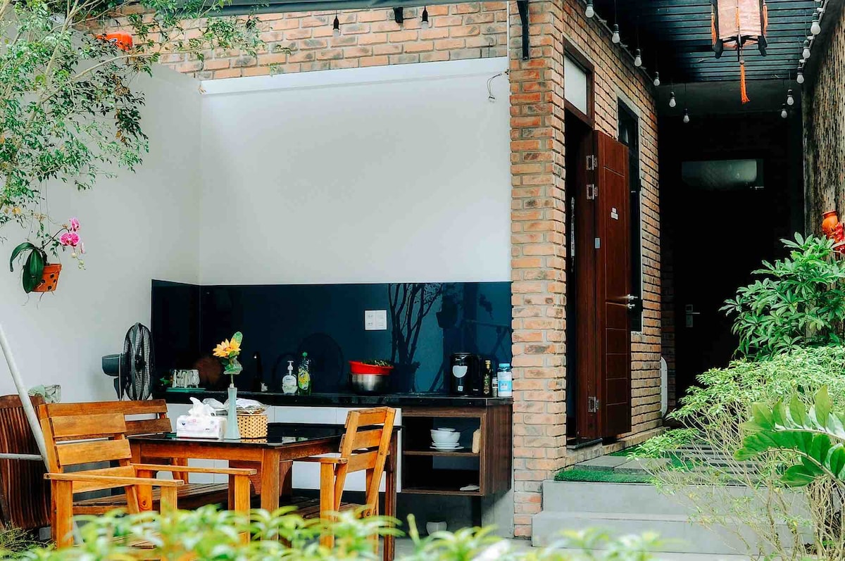An open kitchen area is depicted with wooden chairs and a table, inviting outdoor dining. The kitchen features a dark backsplash and a variety of cookware. Lush greenery surrounds the entrance, which is marked by a wooden door and soft lighting.