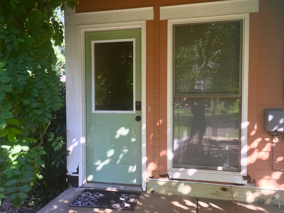 The entrance to the apartment features a light green door with a window, surrounded by a blend of orange siding and lush greenery. A doormat is positioned just in front of the door, while sunlight and shadows play on the surface.