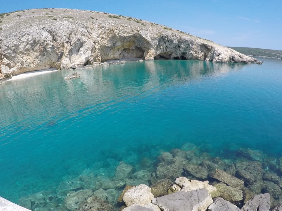 A serene coastal view captures clear turquoise waters gently lapping against rocky shorelines. The landscape features a sheltered cove surrounded by rocky cliffs, with sunlight reflecting off the water's surface, creating a tranquil atmosphere suited for relaxation.