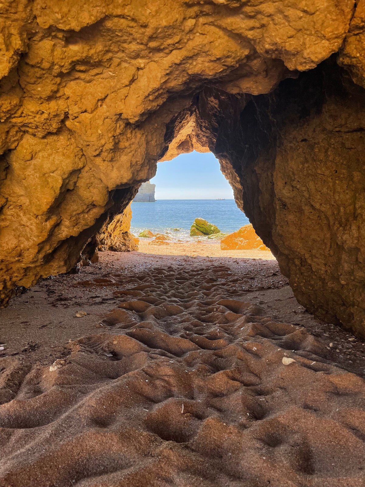 The image captures a cave entrance framed by textured rock walls, leading to a view of the sea beyond. Soft sand is visible at the cave's opening, and sunlight illuminates the scene, creating a contrast between the darker cave interior and the brighter beach outside.
