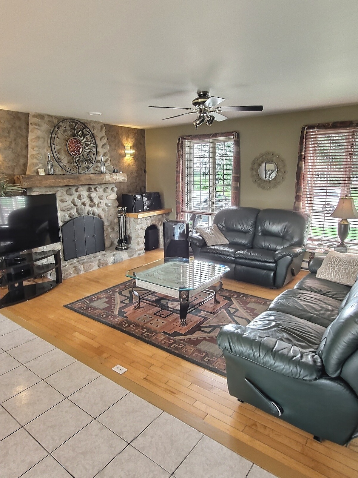 A spacious living area contains leather seating arranged around a glass-topped coffee table. A stone fireplace serves as a focal point, complemented by decorative accents. Large windows allow natural light to filter in, framed by wooden blinds. A television is positioned in the corner.