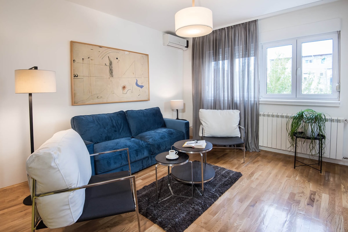 A comfortable living area is furnished with a blue sofa, two armchairs, and a glass coffee table. Natural light enters through a window adorned with sheer curtains. A piece of wall art adds a decorative touch, and a small potted plant enhances the space.