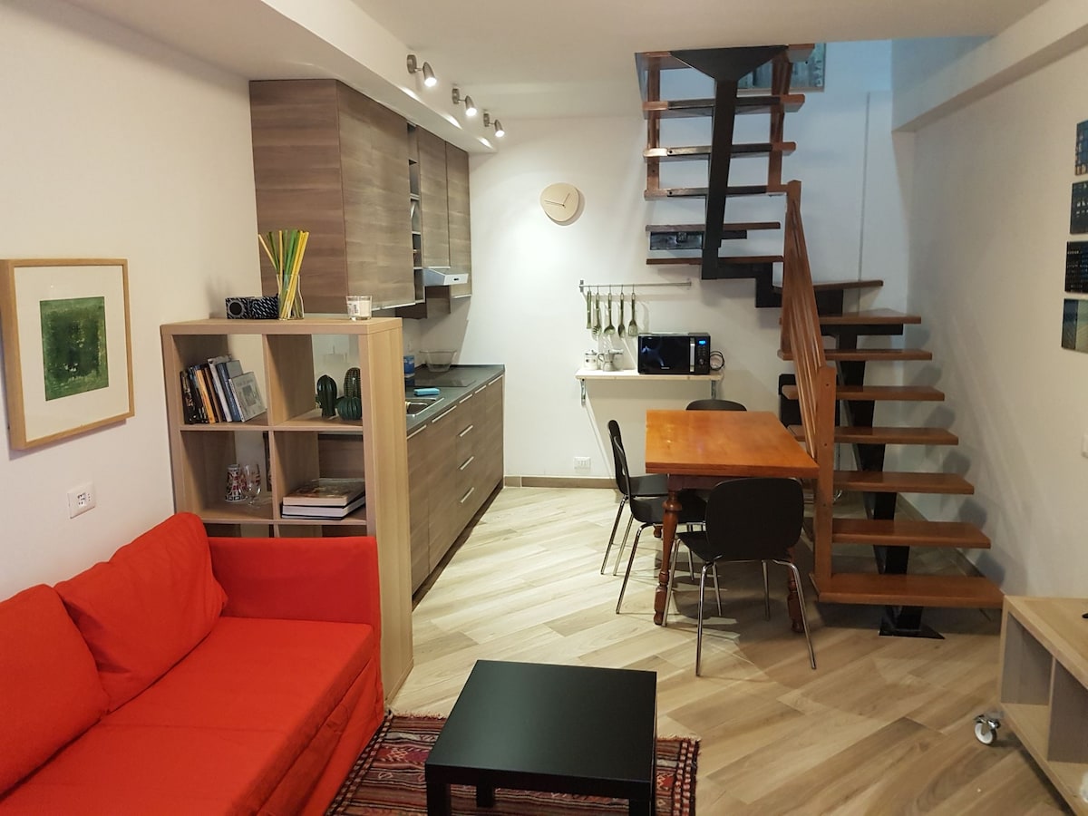 A modern living area features a bright red sofa facing a stylish wooden dining table surrounded by black chairs. Light wood flooring enhances the space, while a staircase ascends to the upper level. A compact kitchen is visible with ample storage and modern appliances.
