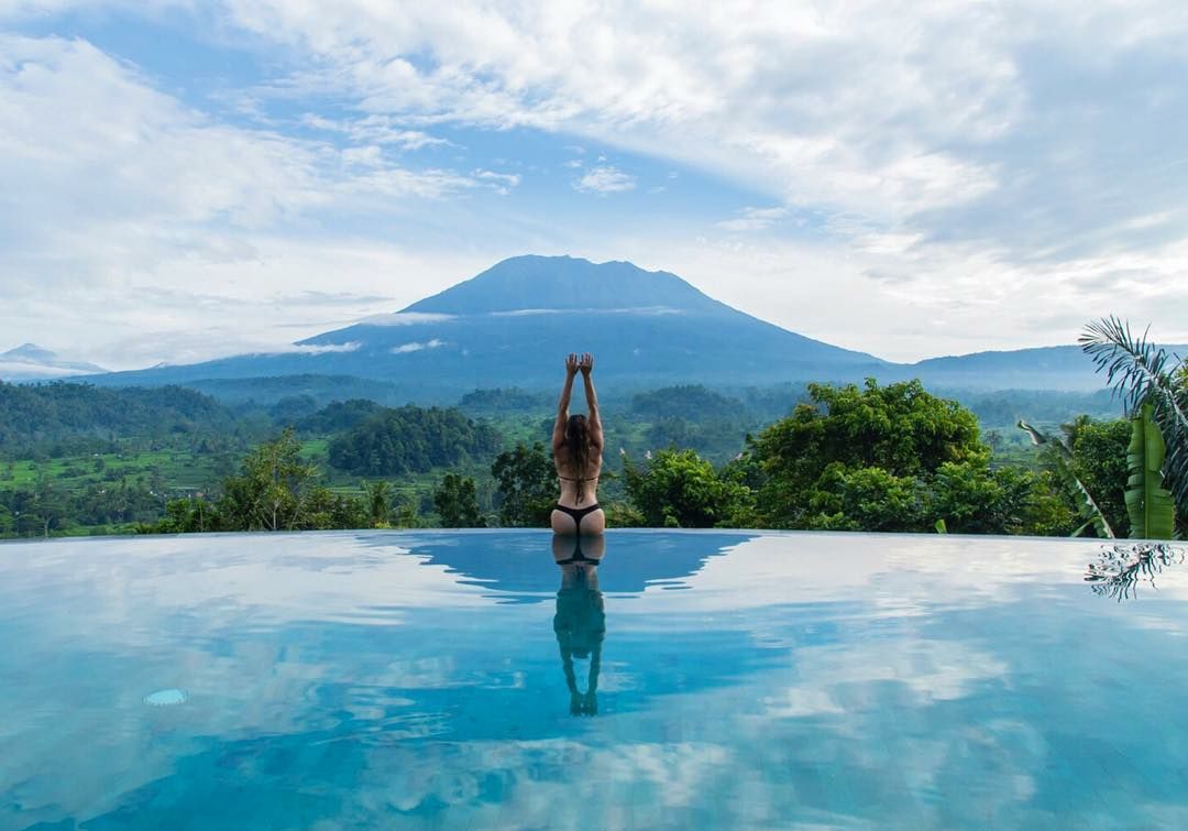 An infinity pool is positioned at the edge of a mountain view, with lush jungle surroundings. A person is seen stretching, creating a connection with the serene landscape. The sky is filled with soft clouds, enhancing the tranquil scene.