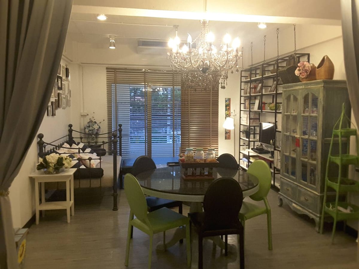 A spacious living area is showcased with a round glass dining table surrounded by green and black chairs. A chandelier hangs from the ceiling, illuminating the space. A stylish bookshelf and a cozy seating area with a bed can be seen beside large windows adorned with blinds.