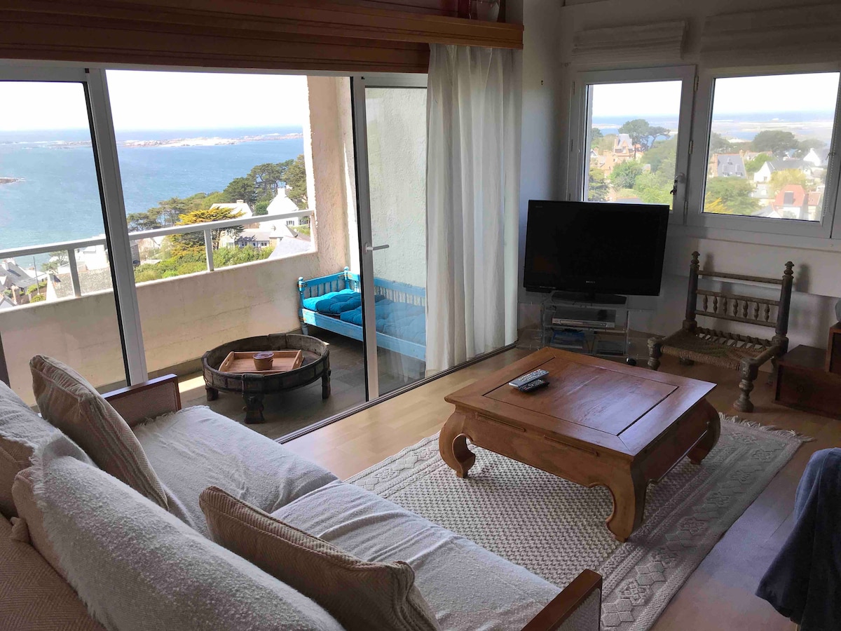 A light-filled living area is presented, featuring a spacious sofa and a wooden coffee table. Floor-to-ceiling glass doors offer stunning views of the coastline. A blue outdoor seating area is visible through the doors, complemented by a decorative area rug on the wooden flooring.