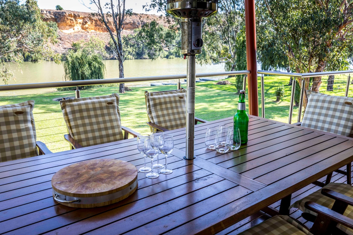 An outdoor dining area features a wooden table surrounded by cushioned chairs with a checkered pattern. A bottle of green beverage and glassware are positioned on the table. Lush green grass and trees are visible in the background, with the river gently flowing nearby.
