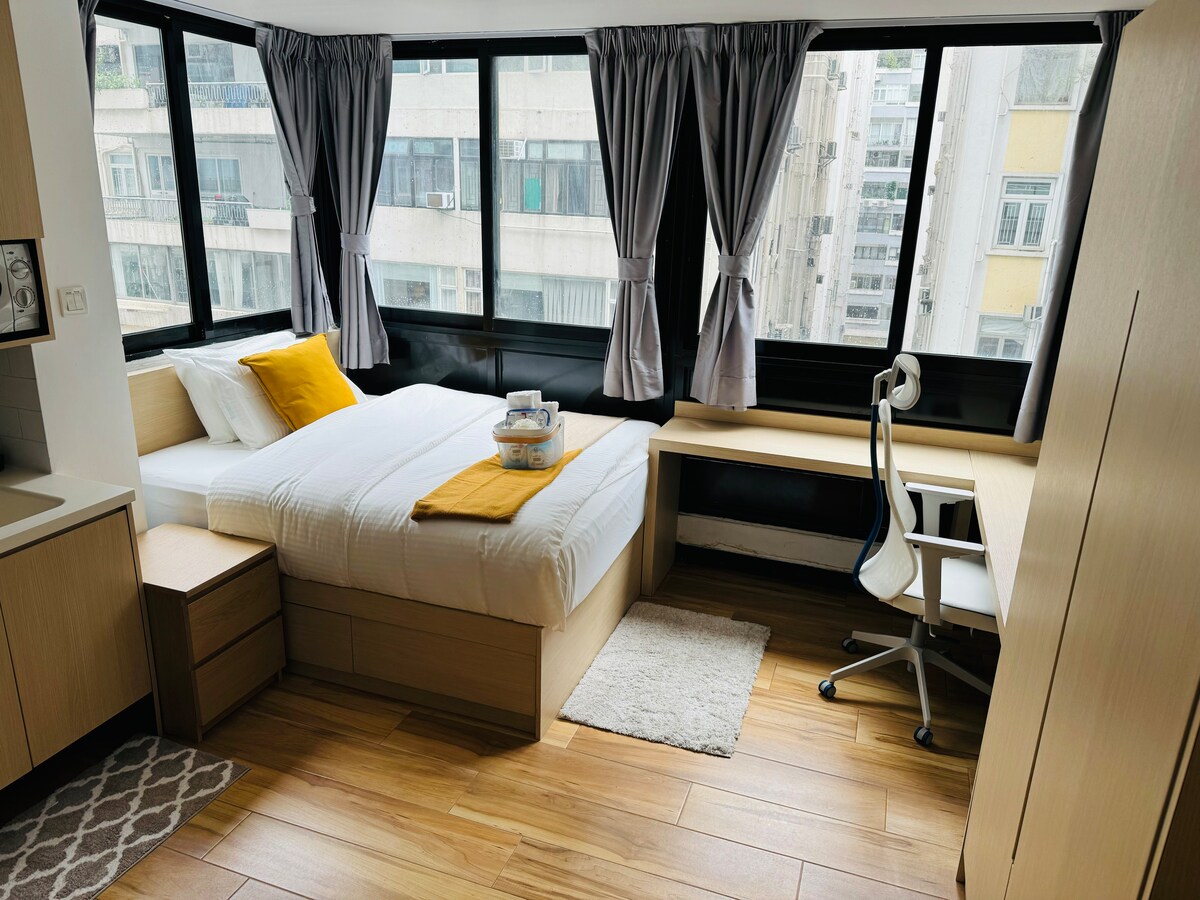 A well-lit bedroom features large windows with sheer curtains that allow natural light to fill the space. A spacious bed is complemented by a yellow accent pillow and a neatly arranged tray. A desk with a comfortable chair and warm wood flooring enhance the inviting atmosphere.