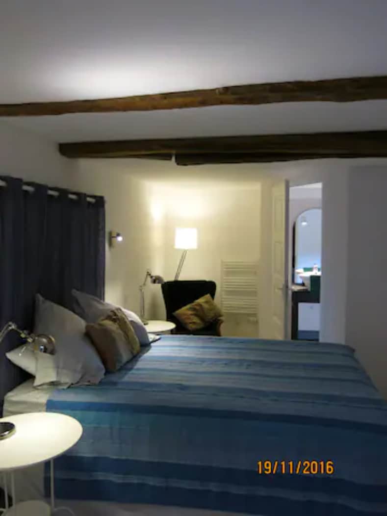 A well-lit sleeping area features a spacious bed with blue and neutral linens. Soft pillows are placed at the head of the bed. A lamp beside the bed provides additional lighting, complemented by a cozy chair in the corner. A doorway leads to a bathroom area.