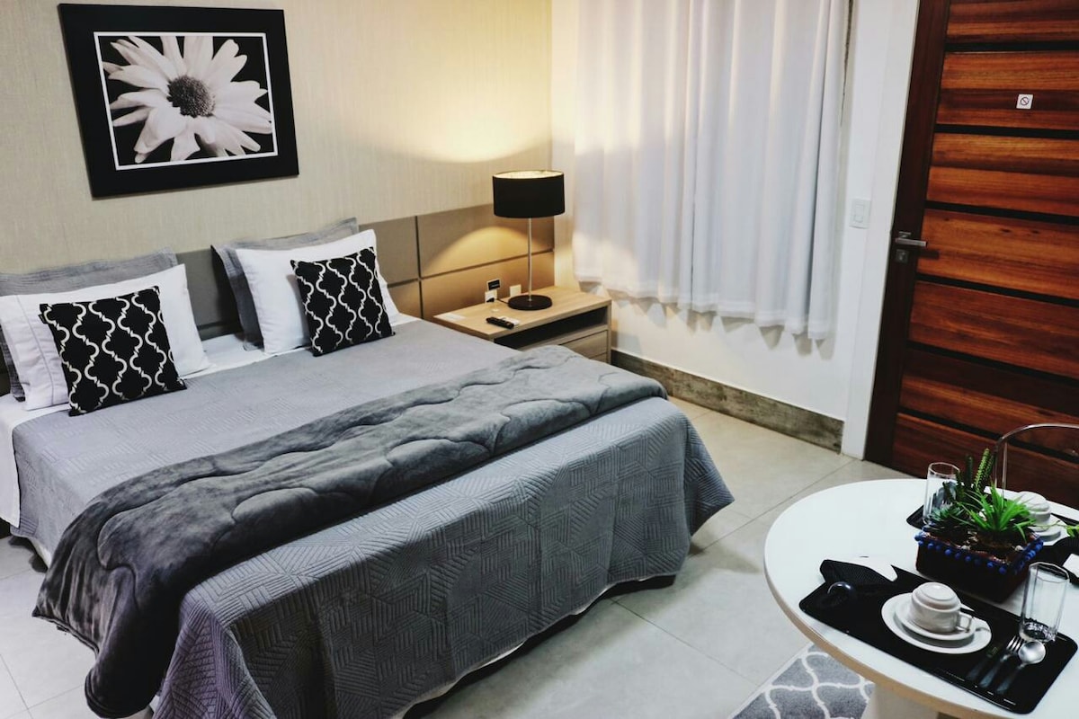 A serene bedroom features a spacious bed with soft gray bedding and decorative pillows. A bedside table is equipped with a lamp and a framed artwork hangs above. Natural light filters through sheer curtains, adding a calm ambiance to the space.