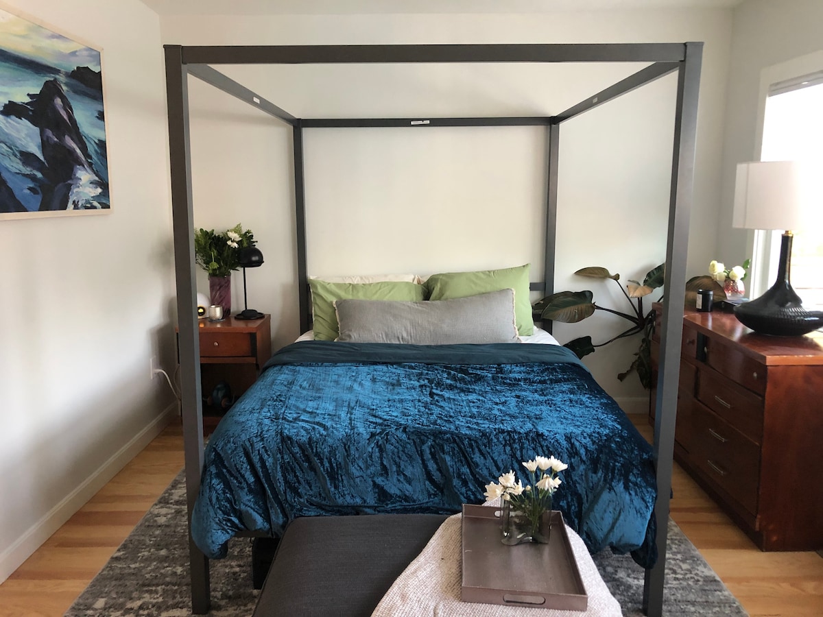 A spacious bedroom is highlighted by a four-poster bed adorned with a teal cover. Complementing the bed are green and gray pillows, a bedside table with a lamp, and a decorative arrangement of plants. Soft natural light enters through a nearby window, enhancing the room's calming ambiance.