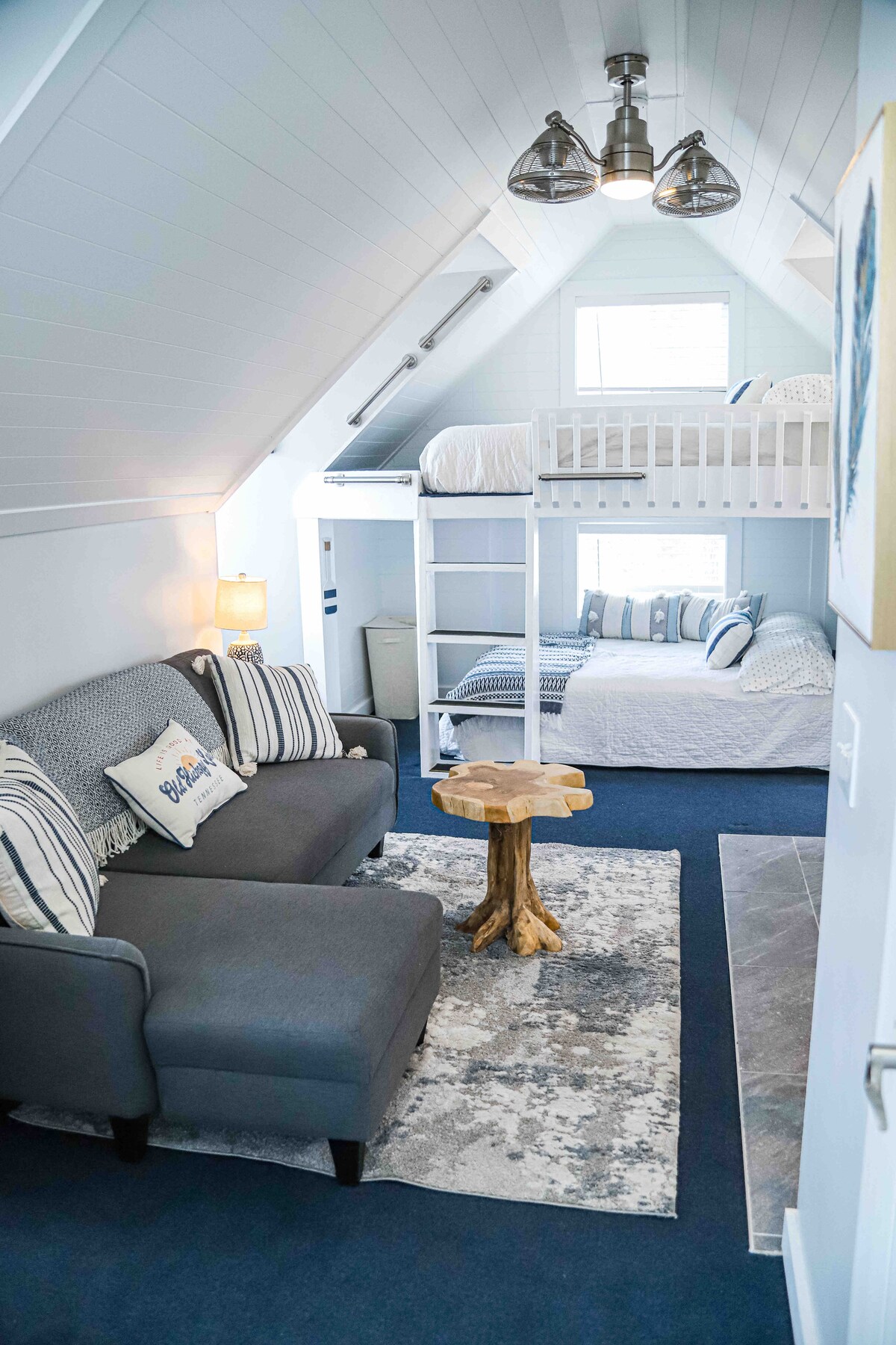 An airy loft space features a cozy seating area with a gray sofa and a tree stump side table. A two-level bed frame stands to the right, showcasing a lower bed with white bedding and an upper bunk with a guardrail, surrounded by bright windows.