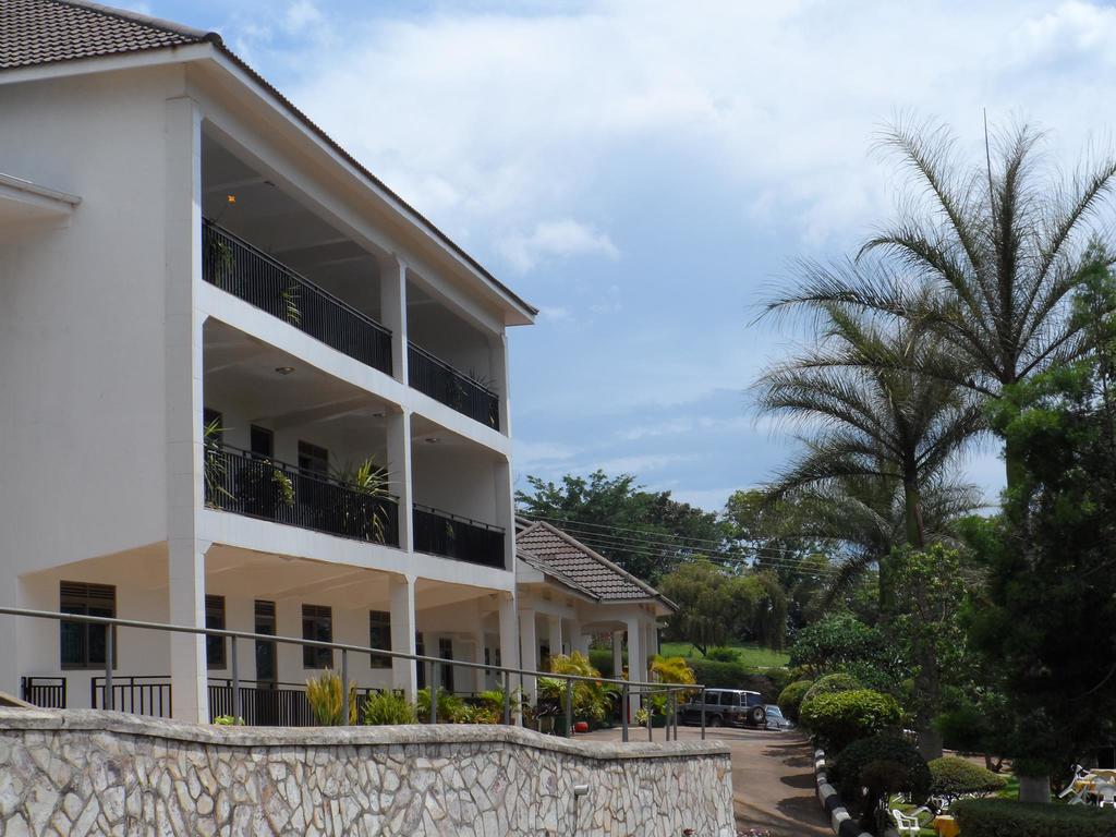 The hotel exterior showcases a two-story building with multiple balconies adorned with greenery. A landscaped garden with various plants and trees is visible in the foreground, while the surrounding area displays a well-maintained driveway and ample parking space.