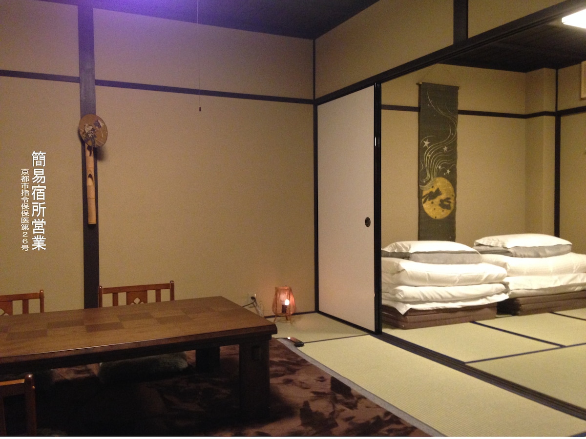 A minimalist room features a low wooden table surrounded by seating, providing an airy feel. Soft, rolled bedding is neatly stacked against the wall. Traditional decor elements, including a hanging artwork and warm lighting, add subtle character to the space, which is adorned with tatami flooring.
