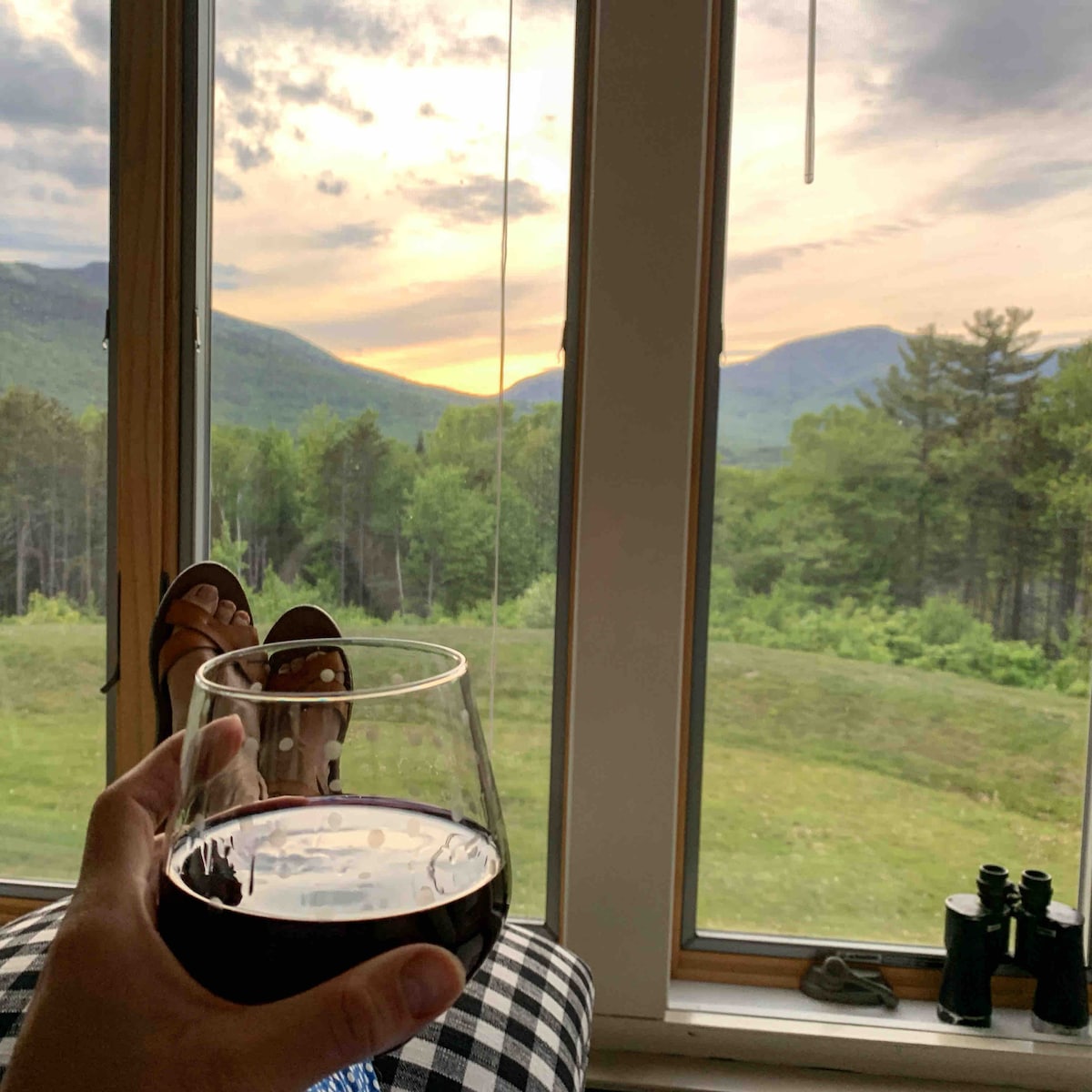 A hand holds a glass of red wine, framed by large windows that offer a view of a sunset over verdant mountains. The foreground features a pair of feet resting on a cozy blanket. Binoculars are positioned nearby, suggesting a serene outdoor experience.